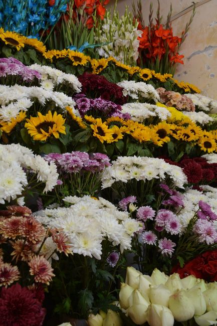 A vibrant display of fresh floral arrangements featuring a variety of flowers. Bright yellow sunflowers with large dark centers are prominently positioned, surrounded by white, purple, pink, and red b