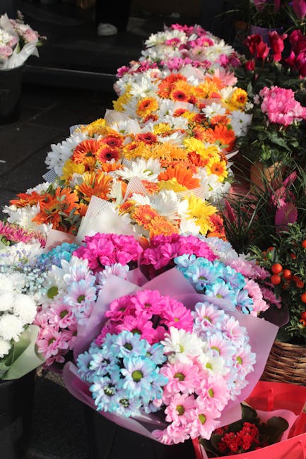A vibrant display of floral bouquets featuring a variety of fresh flowers including pink, white, and blue chrysanthemums, arranged in pastel-colored wrapping paper. The bouquets are placed on a dark s