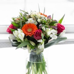 This luxurious bouquet unfolds like a visual poem, shaped with artistry and precision inspired by Bow's creative atmosphere. Creamy-shaded gerbera daisies ring the ensemble with a gentle glow, petals curling softly around golden-orange centres that shine like lanterns at a local festival. The lush, fuchsia-toned roses add depth and drama, their blooms reminiscent of vibrant summer evenings beside local community gardens. Tender sprigs of ivory alstroemeria and petite white filler blossoms emerge among the roses, infusing the arrangement with a lightness and a whisper of fragrance reminiscent of springtime breezes along the Hertford Union Canal. The base is tightly framed by broad, glossy leaves-sweeping, verdant aspidistra folded artfully, lending strength and a grounding sense of tranquility akin to tree-lined Bow streets. Smaller, pointed greens and delicate sprays weave between the central flowers, enhancing volume and contrast. Presented in a clear glass vase, the stems stand proud, echoing the natural beauty of nearby parks. Sunlight caresses the bouquet, making each color vivid but never harsh, gently spotlighting details without overshadowing their subtlety. The whole arrangement sings of optimism, perfect for celebratory gatherings, warm welcomes, or sending encouragement and care to loved ones in Bow. Rooted in local floral tradition, this arrangement is more than decoration-it's a gift of hope, renewal, and heartfelt connection.